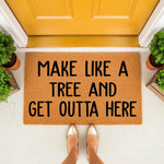 Funny doormat with the text Make Like A Tree And Get Outta Here, a natural coir fiber welcome mat for front porch decor, placed outside a yellow door with potted plants, perfect for a humorous home entrance or housewarming