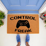 Video game controller doormat with Control Freak text in pixel font, a natural fiber welcome mat for gamers placed at a blue front door, perfect for a gaming room entrance or a fun housewarming gift for a video game enthusiast.