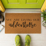 A textured doormat with the phrase "WE ARE LIVING OUR ADVENTURE" printed on it, surrounded by a pair of red sandals and some greenery.
