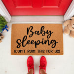 Baby sleeping doormat with funny text, a natural fiber welcome mat for new parents featuring bold black lettering on a tan background, placed at a front door entrance to warn visitors to be quiet and not wake the sleeping infant.