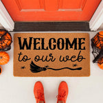 Halloween welcome doormat with Welcome to our web text and a witch broom graphic, featuring a natural coir fiber mat placed on a porch with pumpkins and spider webs for a spooky fall home decor look at a front door entrance.