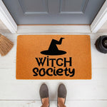 Witch Society Halloween doormat featuring a black hat graphic on a natural coir fiber welcome mat, perfect for fall porch decor and spooky home entryways, shown on a white front porch with a broom and cauldron nearby for seasonal