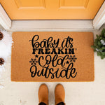 Winter welcome doormat with funny holiday quote Baby It's Freakin Cold Outside, featuring black text and snowflake icons on a natural coir fiber mat placed on a porch with snow and boots for a festive seasonal home entryway decor.