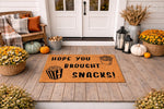 Hope You Brought Snacks doormat, a funny coir welcome mat for front porch decor featuring popcorn and fries illustrations, placed on a rustic wood deck with fall pumpkins and flowers for a cozy seasonal entryway floor mat look.