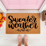 Sweater Weather fall doormat featuring black script text on a natural coir fiber welcome mat, styled on a porch with autumn leaves and a pumpkin for a cozy seasonal home entrance decor look that is perfect for your front door