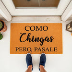 Funny Spanish doormat with black text reading Como Chingas Pero Pasale on a natural fiber welcome mat, placed at a front door entrance with blue sneakers visible, perfect for a humorous home decor porch entry or housewarming.