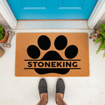Personalized pet paw print doormat featuring a large black paw design on natural coir fiber, perfect as a custom family name welcome mat for dog lovers placed at a front door entrance with a blue door and decorative dog bone