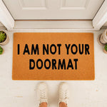 Funny doormat with the text I AM NOT YOUR DOORMAT printed in bold black letters on a natural fiber welcome mat, placed on a porch in front of a white door with a person wearing white sneakers standing on the entryway floor mat.