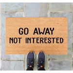 Go Away Not Interested funny doormat made of natural coir fiber, featuring bold black text on a textured brown welcome mat surface, perfect for a front porch entryway to discourage solicitors with a humorous and blunt house decor