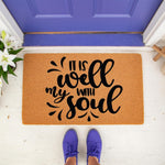 It Is Well With My Soul doormat featuring black script text on a natural coir fiber welcome mat, placed on a porch in front of a purple door with a person wearing purple sneakers standing on the entryway floor mat for home decor.