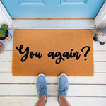 Funny You Again doormat featuring bold black script text on a natural coir fiber welcome mat, placed on a white porch with blue sneakers visible, making this a humorous front door rug for home entryways and housewarming decor.