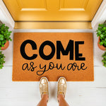 Come As You Are doormat, a natural fiber coir welcome mat with bold black lettering placed on a white porch in front of a yellow door, perfect for a modern home entryway, housewarming decor, or a stylish front door entrance rug.