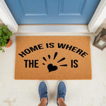 Home Is Where The Heart Is doormat featuring a natural coir fiber texture and black text, placed on a front porch as a stylish welcome mat for your entryway, housewarming decor, or a personalized outdoor floor rug for your home