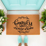 Family Makes a House a Home doormat, a natural coir fiber welcome mat with black script text and leaf accents, placed at a front door entrance with potted flowers and a person wearing teal sneakers standing on the porch floor.