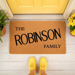Personalized family name coir doormat featuring bold black text on a natural fiber welcome mat, placed on a porch in front of a yellow door with potted plants, creating a stylish custom housewarming entrance decor for any home.