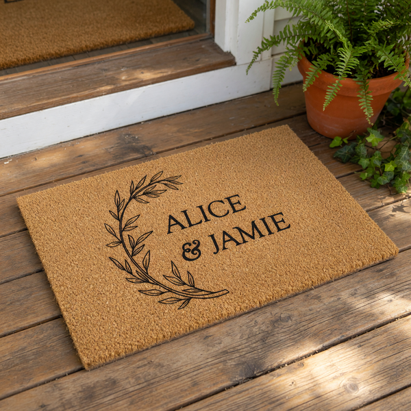 Personalized couple doormat featuring a black botanical wreath design on natural coir fiber, perfect as a custom wedding or housewarming welcome mat for a front porch entrance, showcasing a stylish script and serif typography