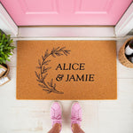 Personalized couple doormat with a natural coir fiber texture, featuring a black botanical wreath design and custom text on a front porch, making a stylish welcome mat for a new home or a housewarming entry decor piece.