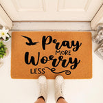 Pray More Worry Less doormat featuring black script text on a natural coir fiber welcome mat, placed at a front door entrance with a person wearing white sneakers standing on it for a stylish and encouraging home decor entryway