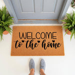 Welcome to the home coir doormat with black script lettering and heart accents, placed on a front porch entryway with potted ferns and gray sneakers, making a stylish outdoor welcome mat for your house entrance or front door