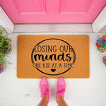 Funny Losing Our Minds One Kid At A Time doormat featuring a circular design on natural coir fiber, perfect as a humorous front porch welcome mat for parents, placed on a white floor in front of a bright pink door with pink