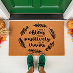 Fall welcome doormat with Smile Often Think Positively Give Thanks text in a black leaf wreath design, featuring a natural coir fiber texture on a front porch with pumpkins and wheat decor for a seasonal autumn home entrance look.