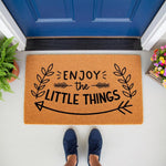 Enjoy the Little Things coir doormat featuring a black arrow and leaf design, placed on a porch in front of a blue door.