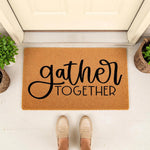 Gather Together coir doormat for front porch entry, featuring a natural fiber welcome mat with black script typography, placed on a white floor with potted plants and a person wearing beige loafers standing in front of a door.