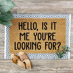 Funny doormat with the text Hello Is It Me You Are Looking For printed on a natural coir fiber mat, layered over a gray and white patterned rug on a wood floor, perfect for a front porch entry or a humorous home decor welcome mat.