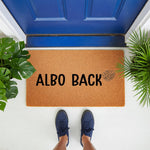 Natural coir doormat with Albo Back text and a monstera leaf illustration, serving as a funny plant lover welcome mat for a front porch entrance, placed on a white floor in front of a blue door with potted tropical house plants