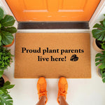Proud plant parents live here doormat featuring a monstera leaf icon, this natural coir fiber welcome mat is a funny housewarming gift for indoor jungle lovers and gardeners placed on a front porch with potted plants and orange