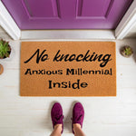 Funny No Knocking Anxious Millennial Inside doormat featuring bold black text on a natural coir fiber welcome mat, placed on a porch in front of a purple door with a person wearing purple sneakers standing on the mat for a