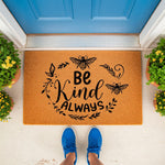 Be Kind Always bee doormat featuring a natural fiber welcome mat with black bee and floral design, placed on a front porch with potted yellow flowers and blue shoes visible, perfect for a farmhouse style home entrance decor.