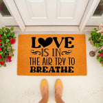 Love Is In The Air Try To Breathe doormat featuring bold black text on a bright orange coir welcome mat, placed on a front porch with potted plants and a person wearing yellow shoes standing in front of the entryway floor decor.