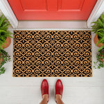 Decorative coir doormat with a black and natural tan floral scroll pattern, placed on a white porch in front of a red door, this durable welcome mat serves as a stylish outdoor floor rug for your home entryway and front porch