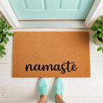 Namaste coir doormat for front porch entry, featuring a natural fiber welcome mat with black script text, placed on a white floor in front of a light blue door with green potted plants for a stylish home decor entrance look.