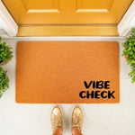 Vibe Check doormat for front porch entry, featuring a natural coir fiber welcome mat with bold black text, placed on a clean entryway floor with potted plants and gold sneakers visible in this stylish home decor product photo.