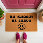 Be Groovy Or Leave doormat with a retro smiley face design, featuring a natural coir welcome mat on a white porch floor in front of a pink door, perfect for a fun front door entrance or a housewarming decor piece for your home.