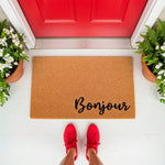 Bonjour coir doormat for front porch entry, featuring a natural fiber welcome mat with black script text placed on a white deck in front of a bright red door with potted plants, creating a stylish outdoor home decor entrance look.