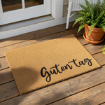 Guten Tag coir doormat featuring black cursive text on a natural fiber welcome mat, placed on a wood porch floor near a potted fern, perfect for a stylish front door entrance or a housewarming gift for a new home owner.