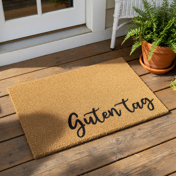 Guten Tag coir doormat featuring black cursive text on a natural fiber welcome mat, placed on a wood porch floor near a potted fern, perfect for a stylish front door entrance or a housewarming gift for a new home owner.