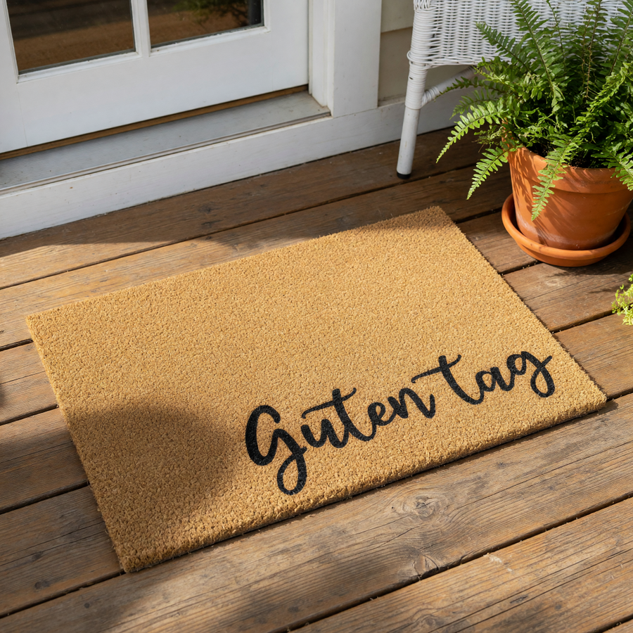 Guten Tag coir doormat featuring black cursive text on a natural fiber welcome mat, placed on a wood porch floor near a potted fern, perfect for a stylish front door entrance or a housewarming gift for a new home owner.