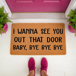Funny coir doormat with black text reading I wanna see you out that door baby bye bye bye, placed on a white porch in front of a pink door, making this a humorous welcome mat for home entryways and a unique housewarming decor