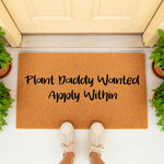 Plant Daddy Wanted Apply Within coir doormat for houseplant lovers, featuring funny text on a natural fiber welcome mat placed at a front door entrance with potted plants and a person wearing casual sneakers standing on the rug.