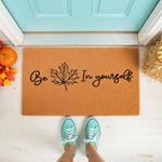 Fall leaf welcome doormat with Be Leaf In Yourself text, a natural coir fiber outdoor rug for autumn porch decor, featuring a black maple leaf design on a tan mat placed in front of a bright blue door with seasonal pumpkins and