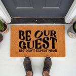 Funny Be Our Guest But Don't Expect Much doormat for front porch, featuring bold black text on a natural coir fiber welcome mat, creating a humorous entryway decor piece for your home entrance that greets visitors with a witty