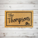 Personalized family name coir doormat with a black border and paw print design, featuring custom text on a natural fiber welcome mat for pet lovers, displayed on a rustic white wood background for a farmhouse entryway decor look.