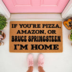 Funny welcome doormat with bold black text on a natural fiber rug, featuring a humorous message about pizza and music deliveries for your front porch decor, placed on a white floor in front of a pink door with potted plants and a