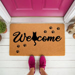 Cat welcome doormat with black paw prints and a feline silhouette on a natural coir fiber mat, placed on a white porch in front of a bright pink door for a cute pet lover home entrance decor and outdoor floor rug entryway
