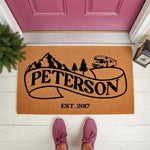 Personalized camping doormat with a mountain, pine tree, and motorhome design on a natural coir welcome mat, featuring a custom family name and established year for a unique outdoor home entrance decor piece for RV enthusiasts.