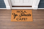 Funny coffee lover doormat with Spanish text Hola Ya Tomaste Cafe printed in black on a natural coir fiber welcome mat, placed on a wood porch in front of a white door for a stylish and inviting home entryway decor look.
